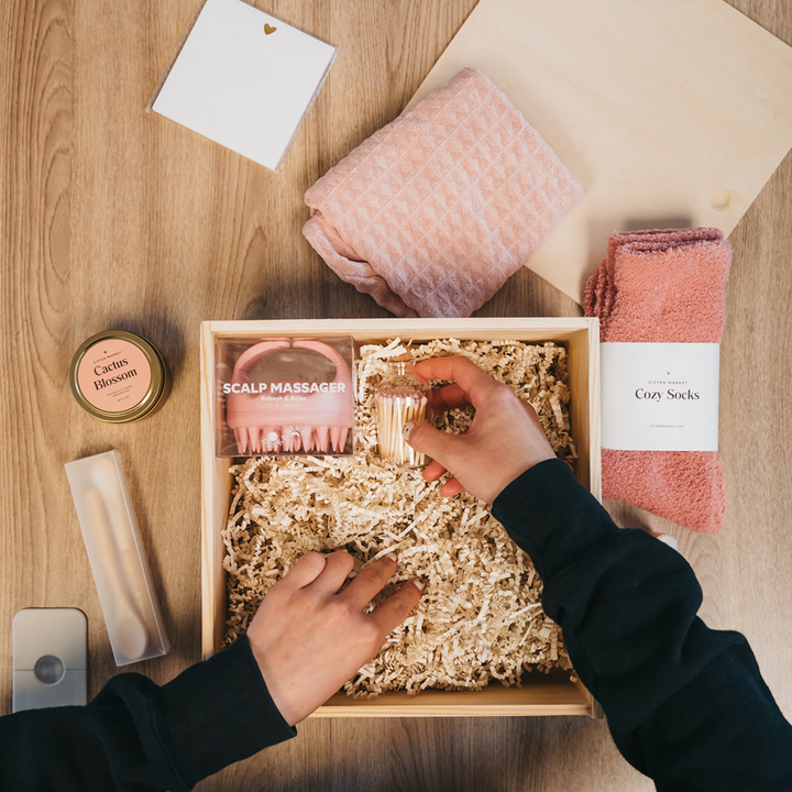 Person opening a box of self-care items including a scalp massager, candle, and socks on a wooden surface.