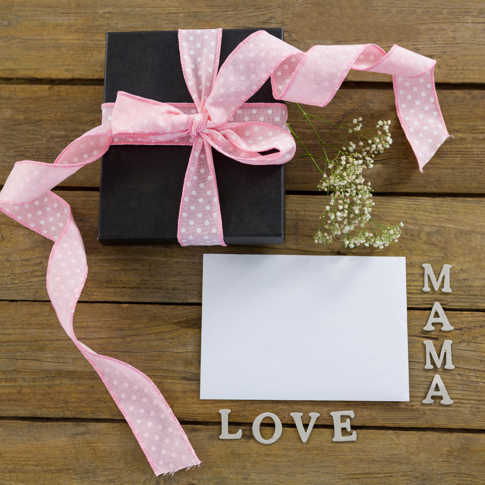 Gift box with pink ribbon, white card, LOVE MAMA letters, and small flowers on wood table