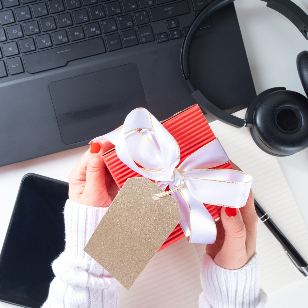 Red gift box with white ribbon and gold tag held above laptop, notebook, and headphones
