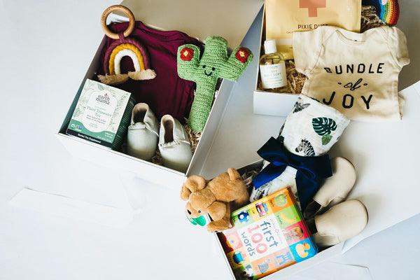White baby gift boxes with plush toys, onesies, baby shoes, board book, and organic snacks