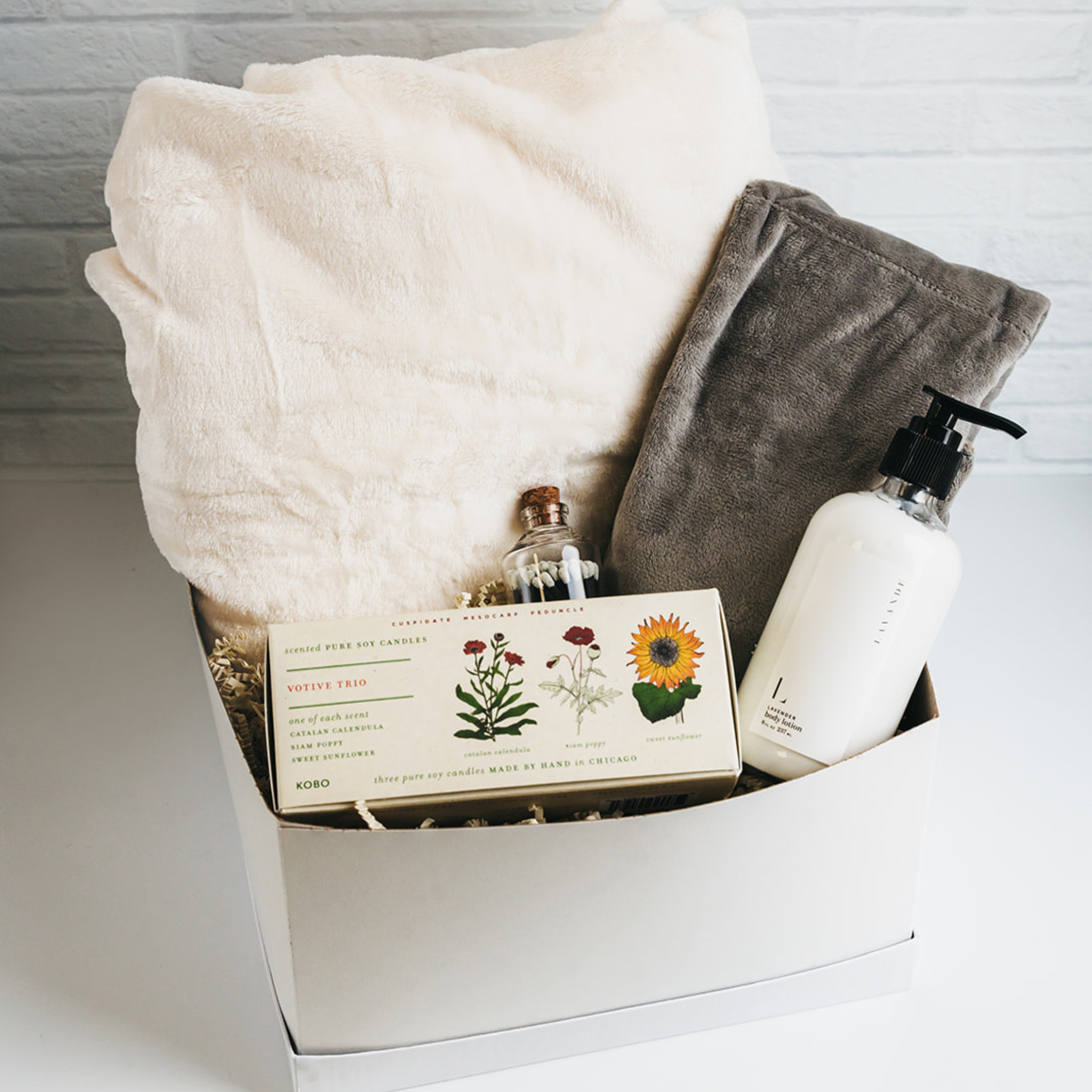 White gift box with plush blanket, grey throw, votive candles, glass jar, and hand lotion