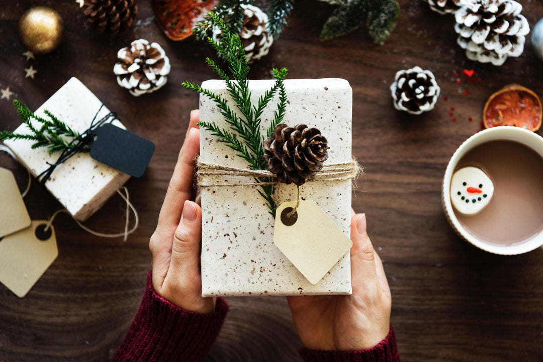Holiday gift box with pine sprig and pinecone, surrounded by festive treats and hot cocoa
