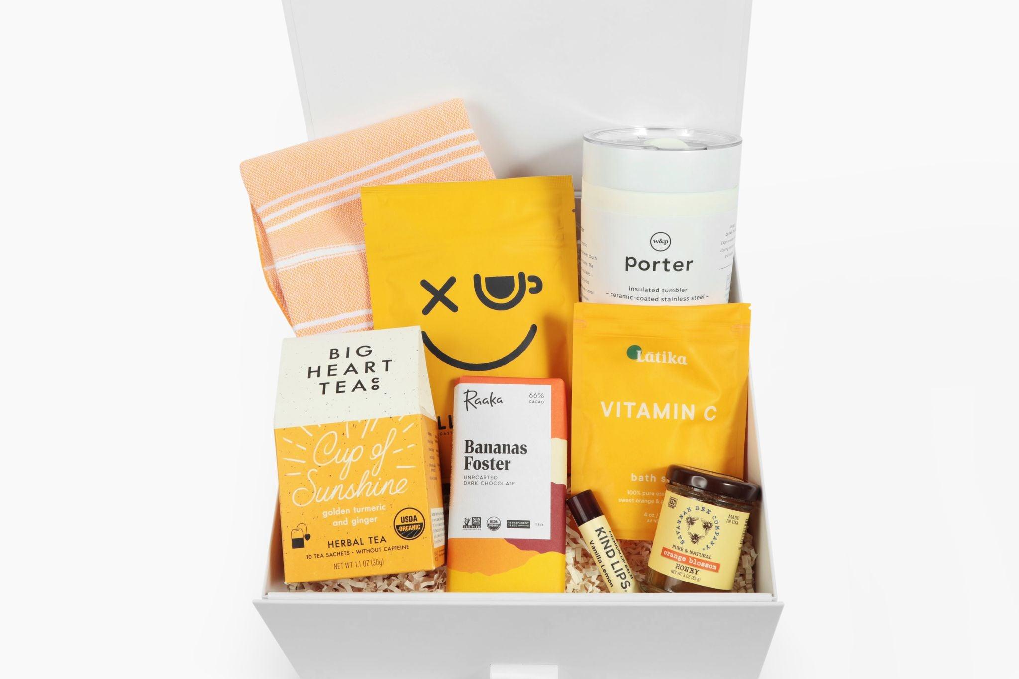 White gift box with tumbler, herbal tea, chocolate bar, honey jar, lip balm, bath soak, snack pouch, and towel