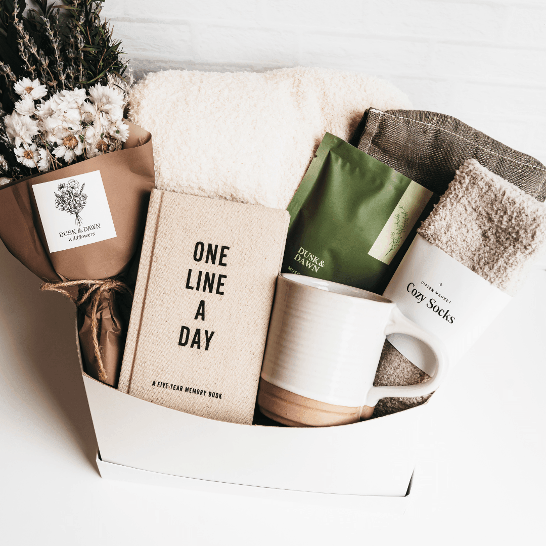 Gift box with wildflowers, memory book, cozy socks, ceramic mug, tea, and blanket.