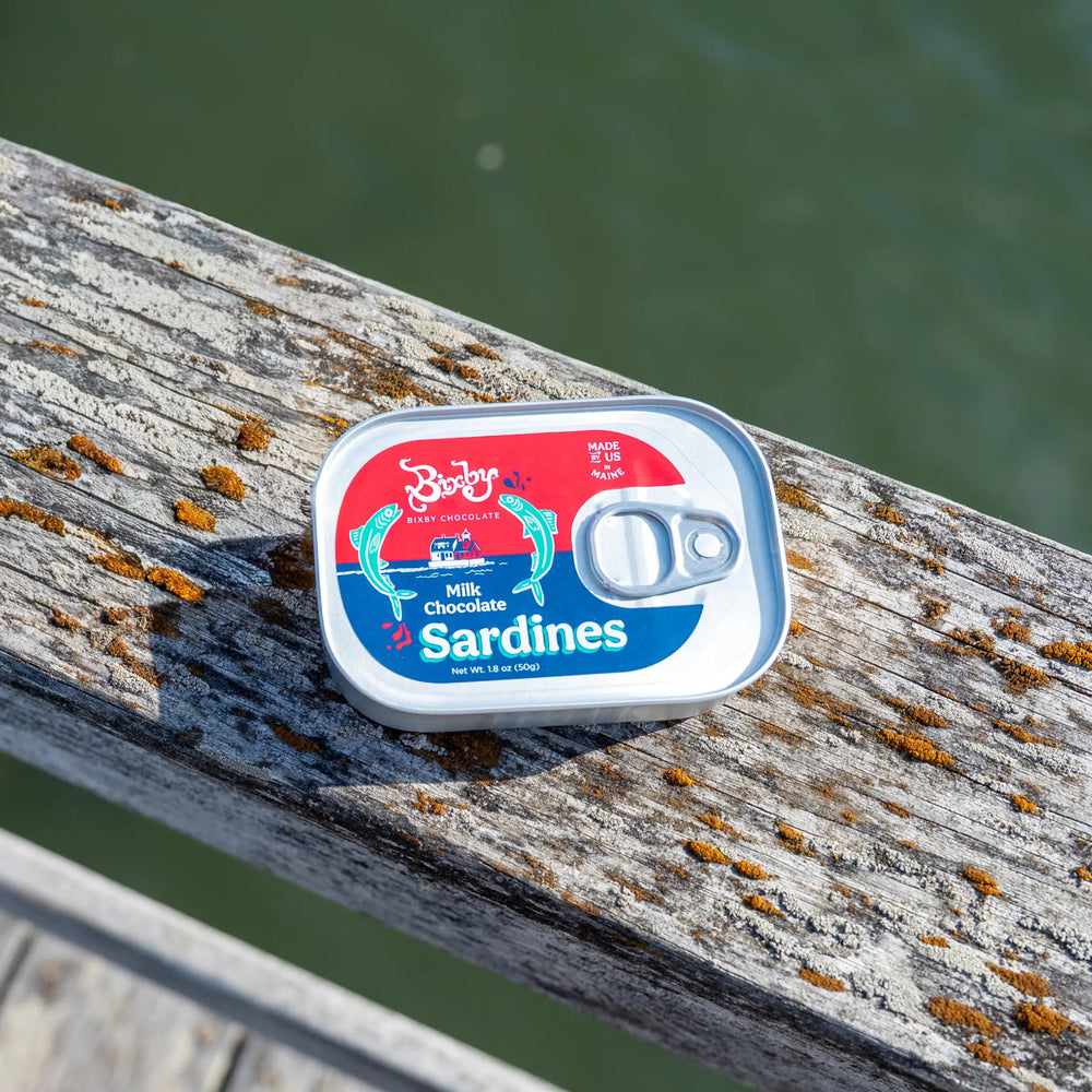 Bixby Milk Chocolate Sardines tin resting on a weathered wooden dock railing above the water