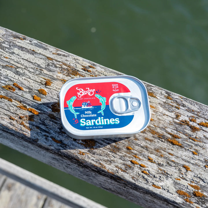 Bixby Milk Chocolate Sardines tin resting on a weathered wooden dock railing above the water