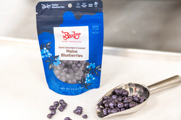 Bag of Bixby chocolate covered blueberries beside a metal scoop of berries