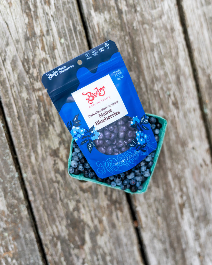 Bixby chocolate covered blueberries bag tucked into a basket of fresh blueberries on weathered wood