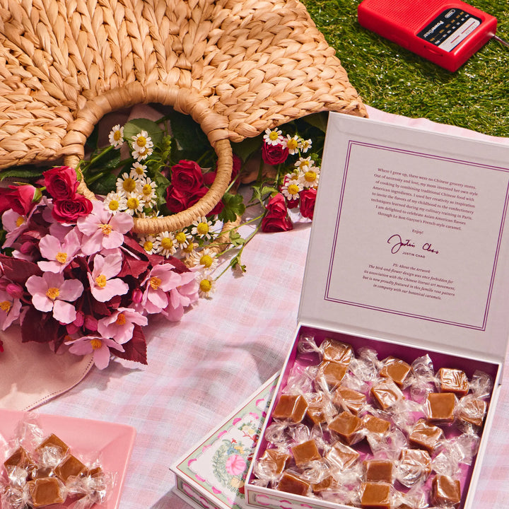 Open Strawberry Sakura caramel box beside a woven basket and pink flowers on a picnic blanket