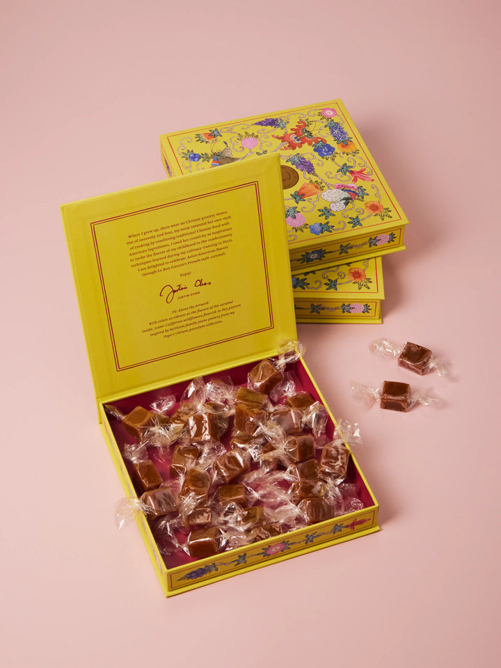 Open yellow floral gift box filled with individually wrapped caramels, on a pink background