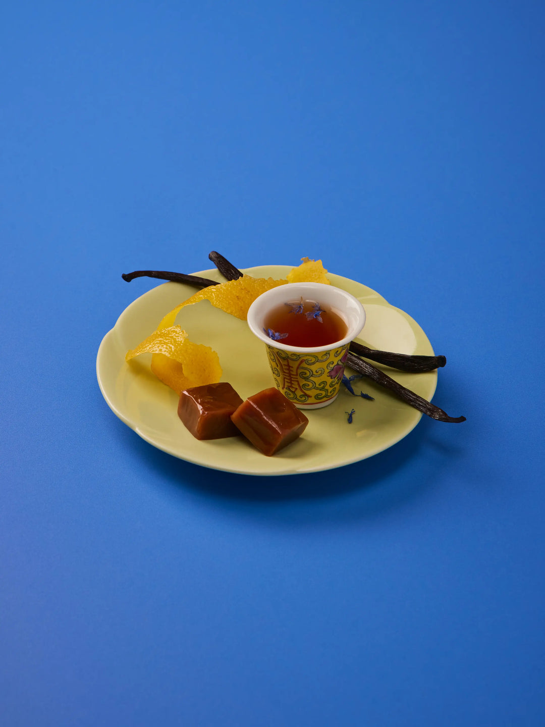 Yellow plate with caramels, a cup of tea, vanilla beans and orange peel on a blue background