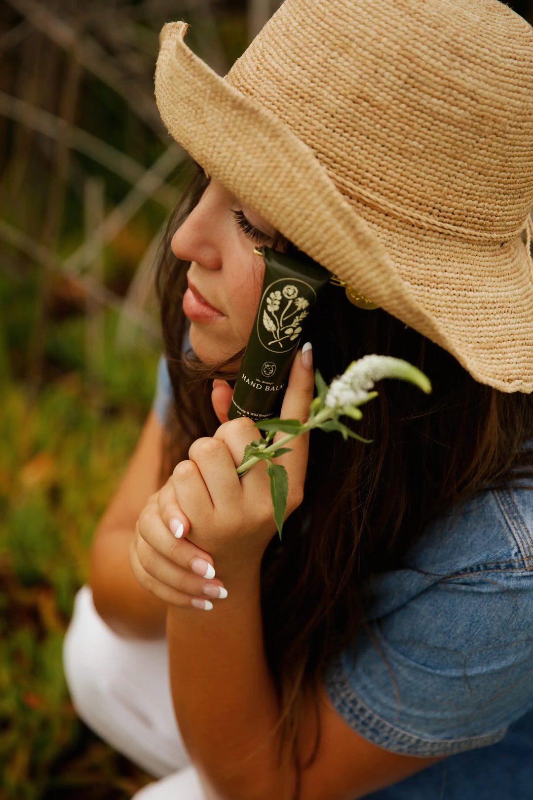 Woman in a straw hat holding the black hand balm tube beside a sprig of wildflowers
