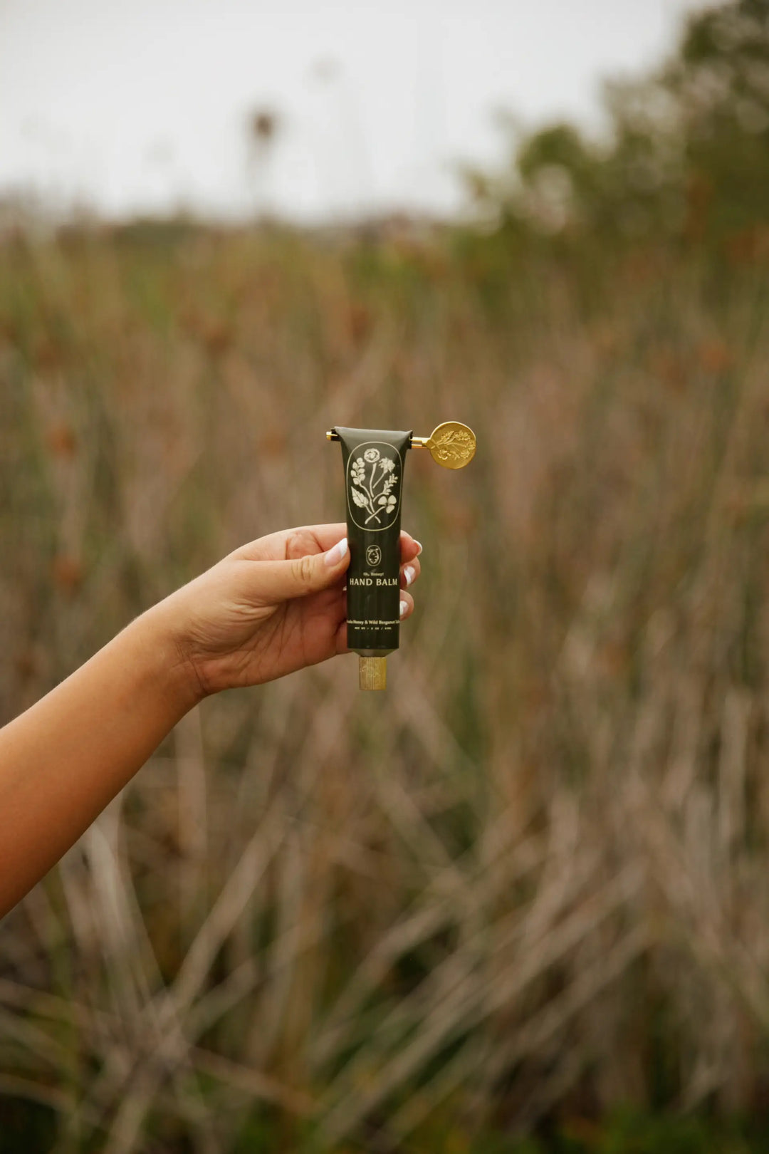 Hand holding the black hand balm tube up against a field of tall grass