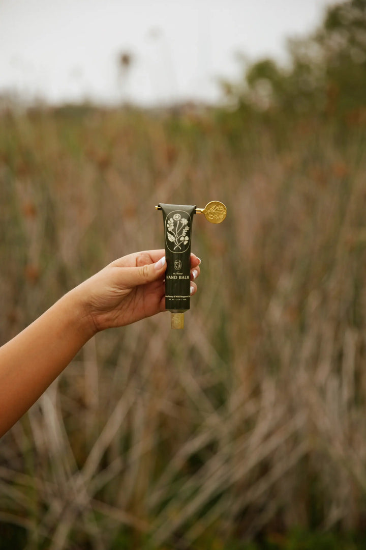 Hand holding the black hand balm tube up against a field of tall grass