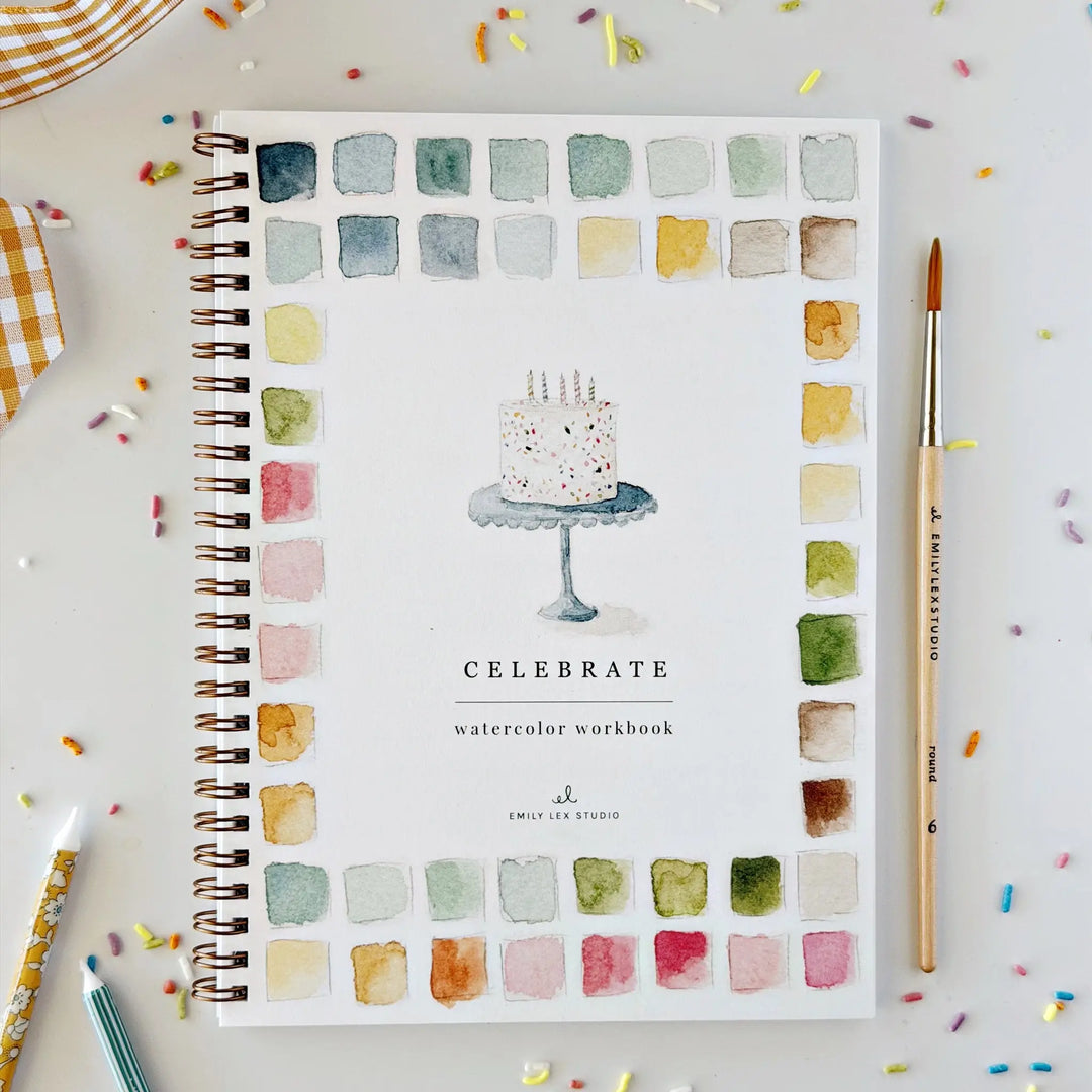 Celebrate Watercolor Workbook cover with a painted birthday cake and watercolor swatch border, beside a paintbrush on a table with sprinkles