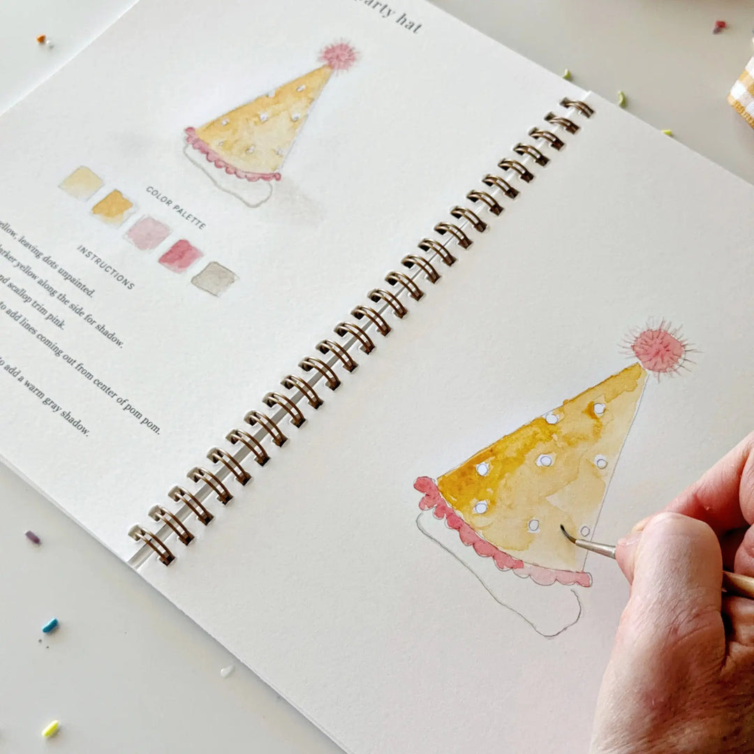 Hand painting a yellow party hat with a pink pom pom on an open page of the workbook