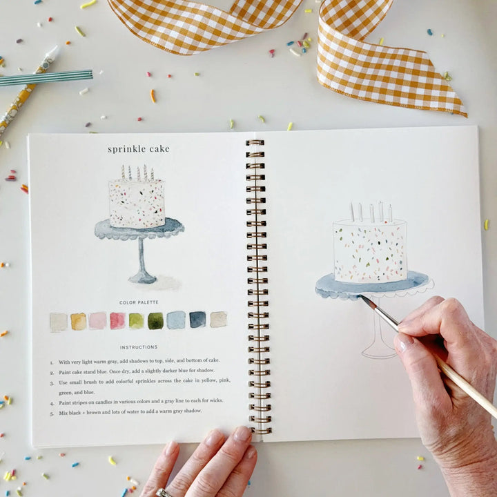 Open spiral workbook with the sprinkle cake page, its color palette and instructions, and a hand painting the cake on the facing page