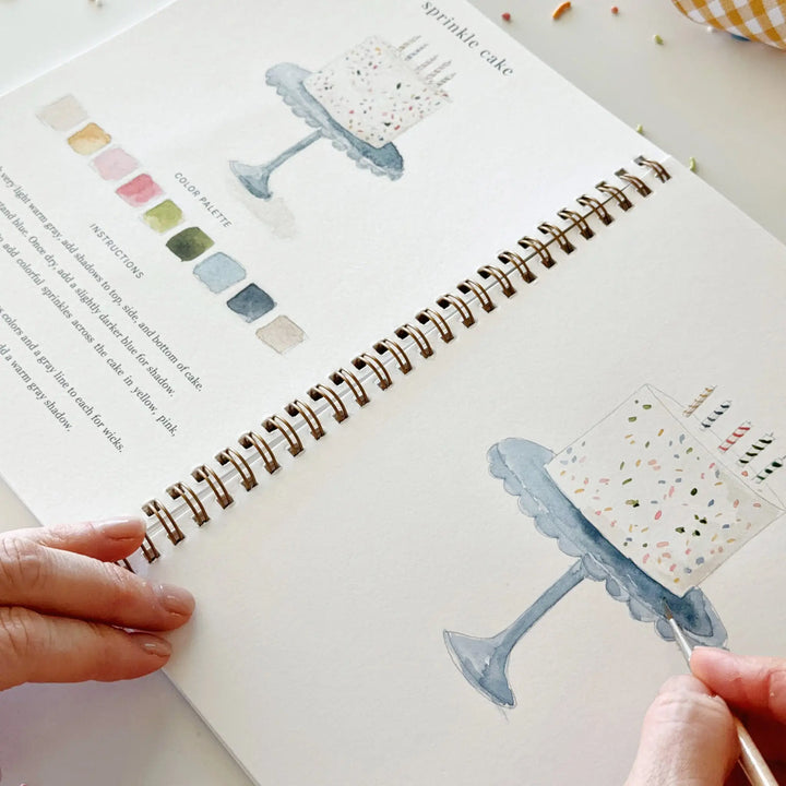 Close view of a hand painting a sprinkle cake on a blue cake stand in the workbook