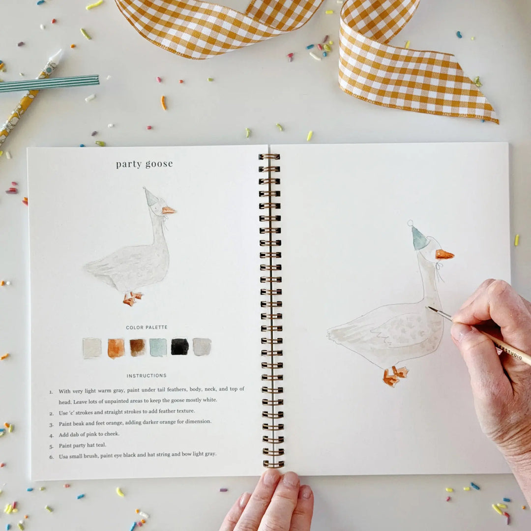 Hand adding details to a goose in a party hat on the facing page of the open workbook