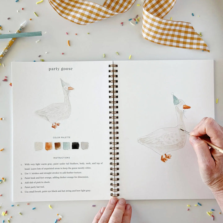 Hand adding details to a goose in a party hat on the facing page of the open workbook
