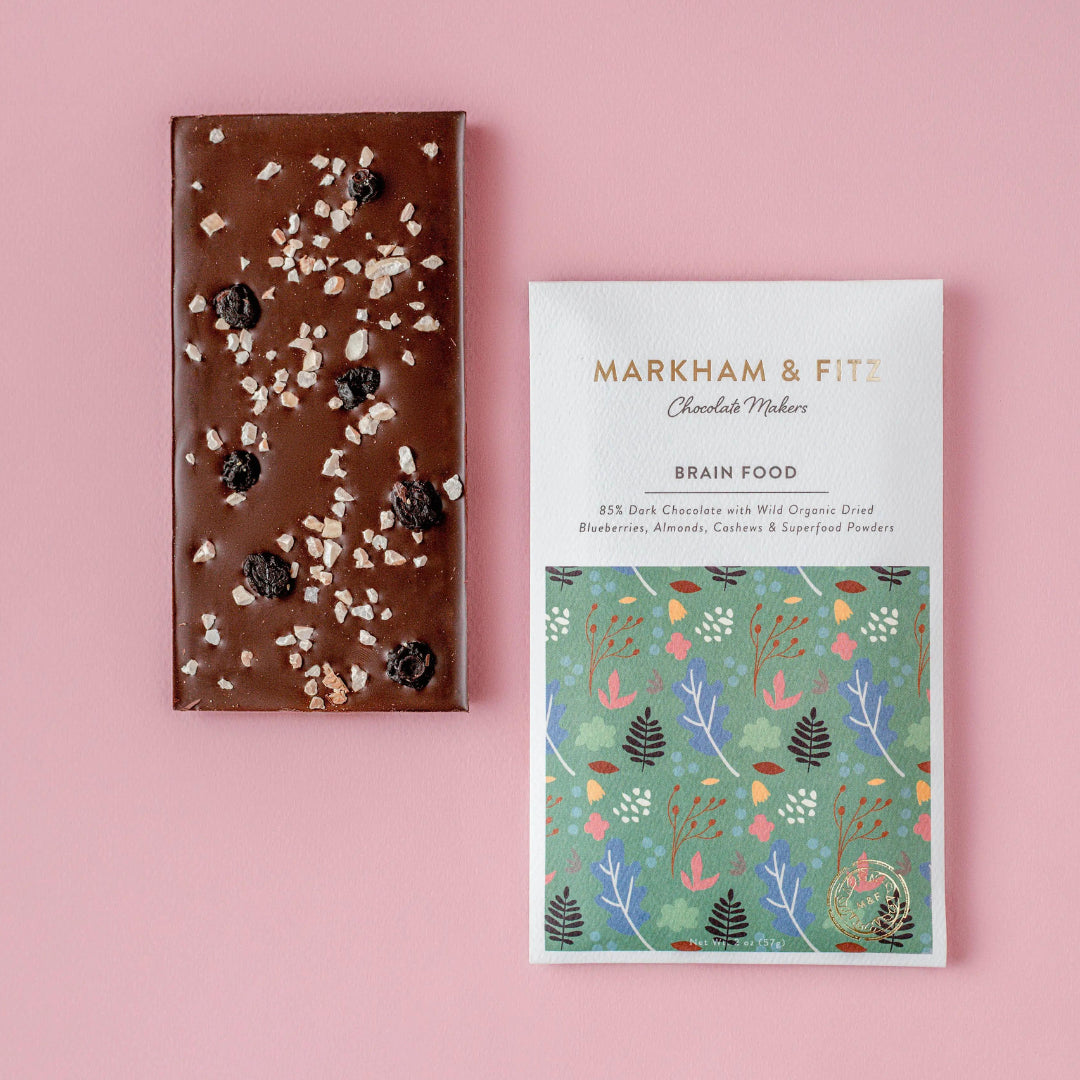 Unwrapped Brain Food dark chocolate bar with dried blueberries and chopped almonds and cashews, next to its sleeve on a pink background