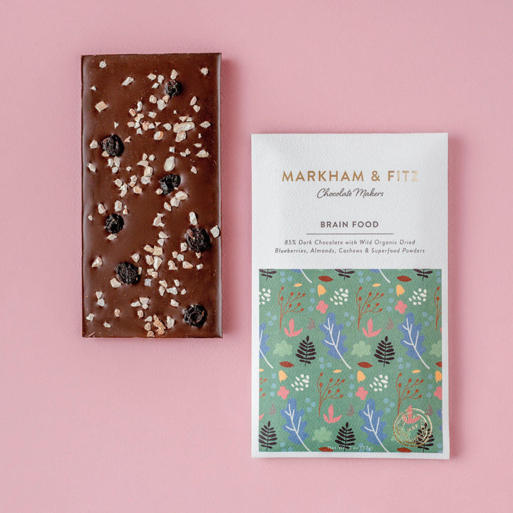 Unwrapped Brain Food dark chocolate bar with dried blueberries and chopped almonds and cashews, next to its sleeve on a pink background