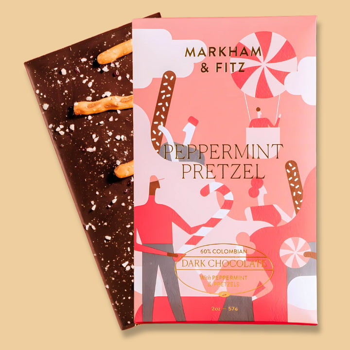 Peppermint Pretzel bar in its coral holiday sleeve, with the dark chocolate bar behind it topped with pretzel pieces and crushed peppermint
