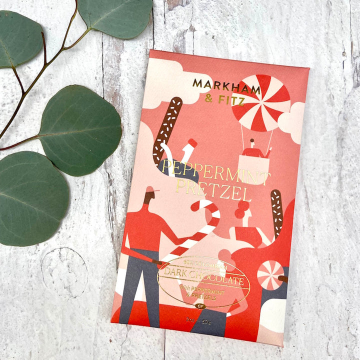 Peppermint Pretzel bar in its illustrated holiday sleeve on a white wood surface with eucalyptus leaves