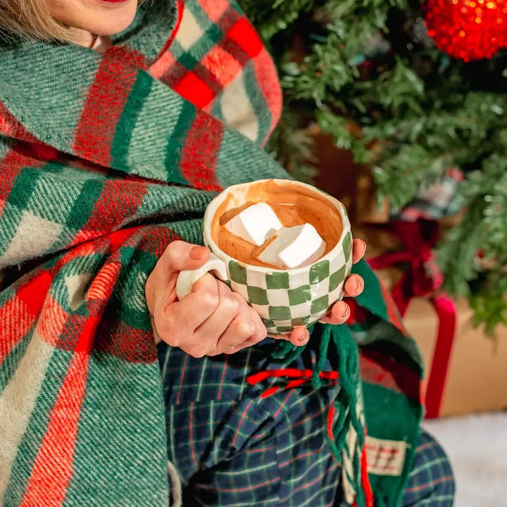 Mug of hot chocolate topped with marshmallows, held in front of a Christmas tree