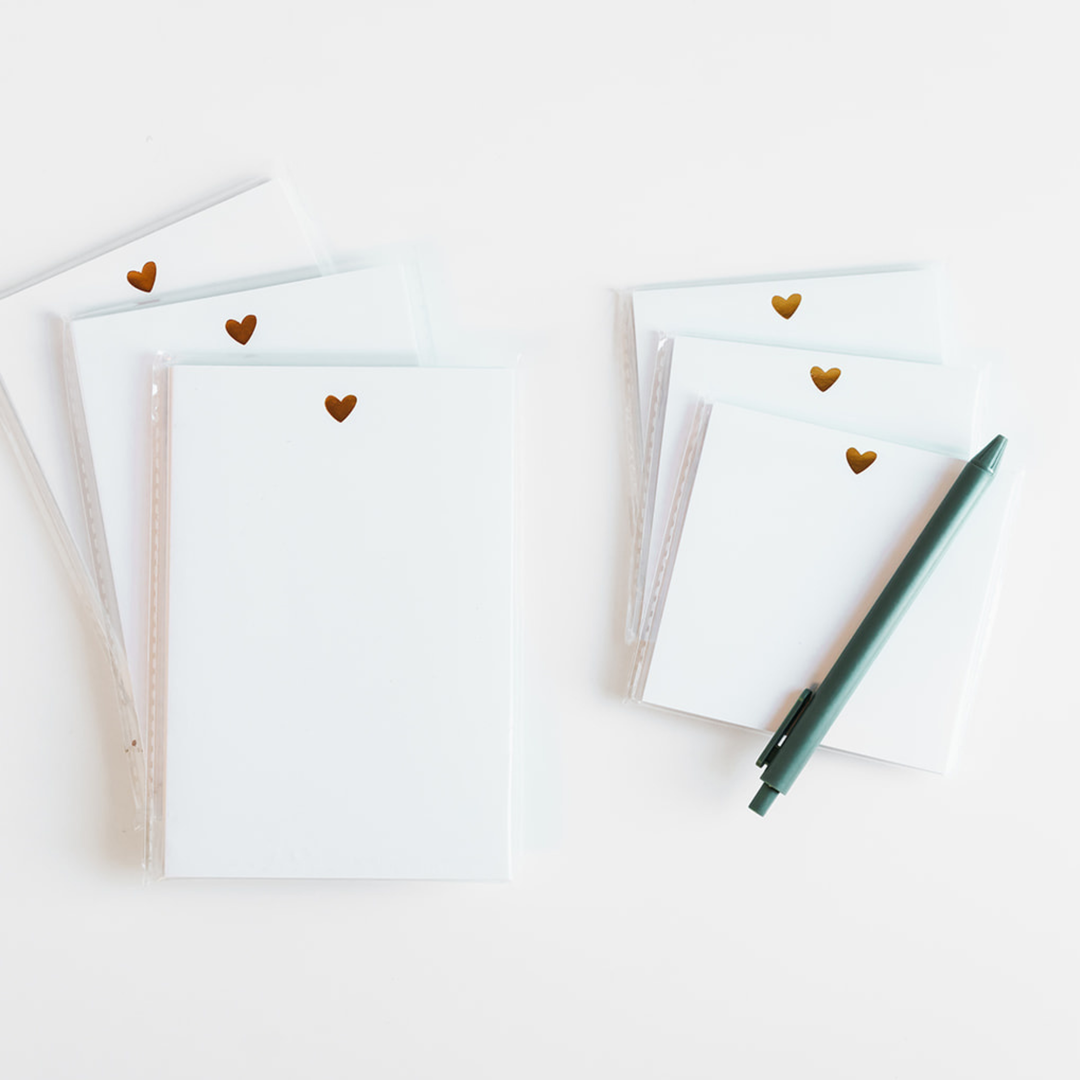 Stacks of white notepads with gold hearts and a green pen on a light background