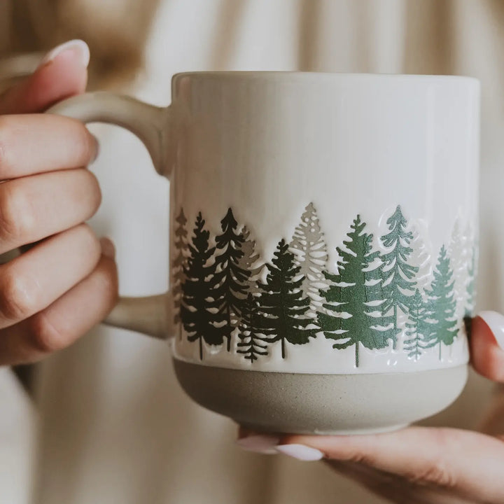 Hands holding the cream speckled stoneware mug, close enough to show the green and taupe tree band and the unglazed base