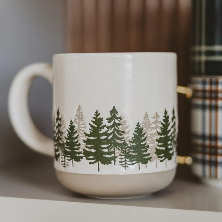The cream stoneware mug on a shelf, green tree design facing forward, next to a plaid mug