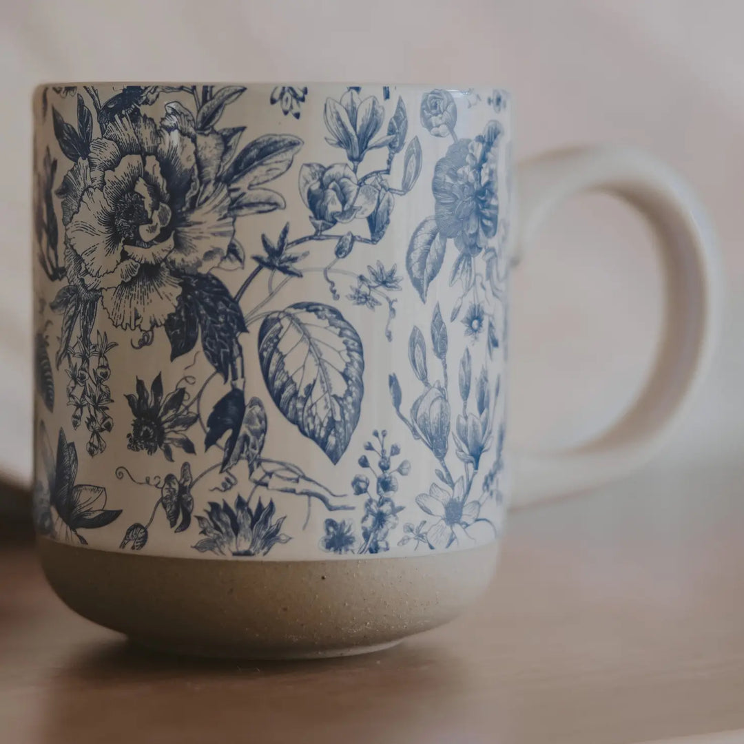 Cream stoneware mug with a blue floral Delft pattern, speckled natural clay band at the base, on a light surface.
