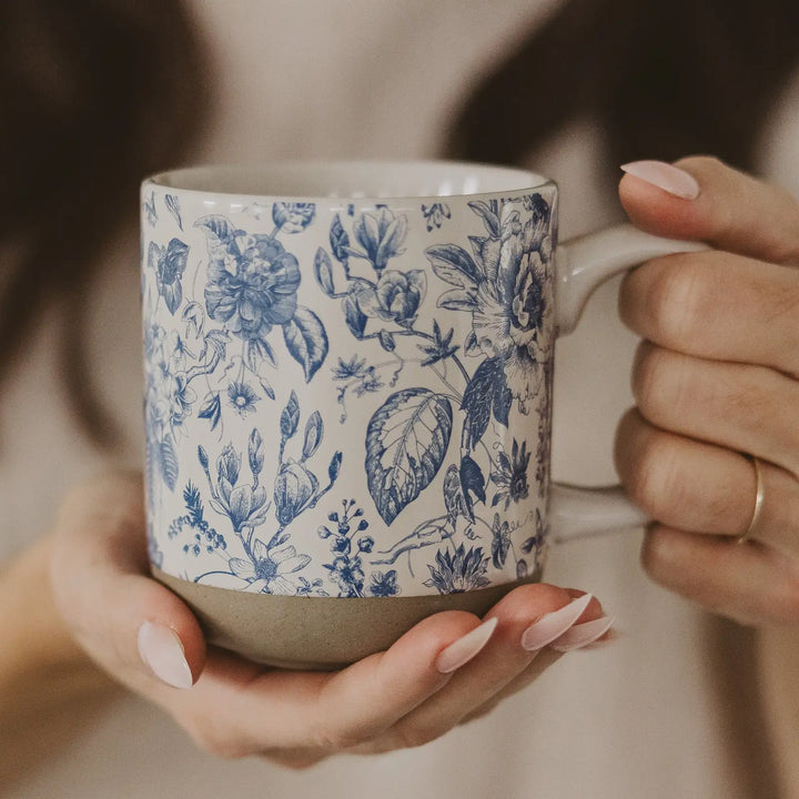 Hands holding the blue floral stoneware mug, showing its full height and the unglazed clay base.