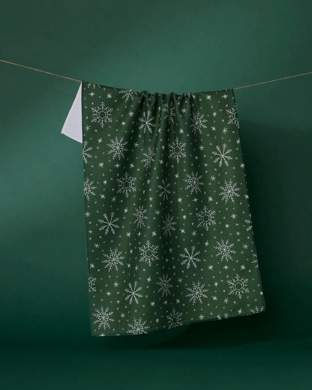 Forest green Firlight tea towel with a white snowflake print, hanging from a line against a green backdrop, its plain white reverse showing at one corner