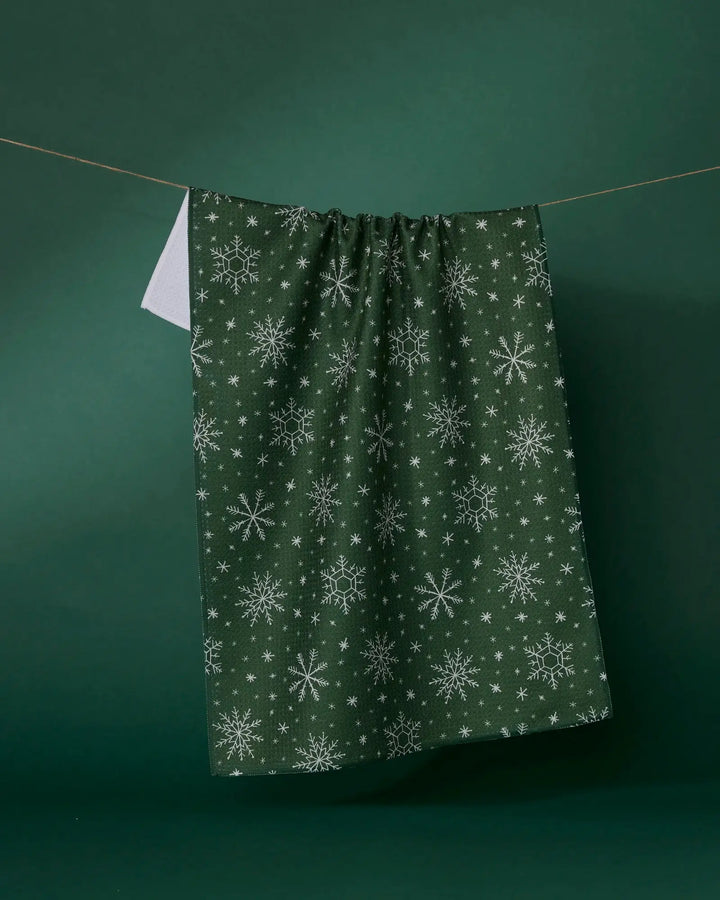 Forest green Firlight tea towel with a white snowflake print, hanging from a line against a green backdrop, its plain white reverse showing at one corner