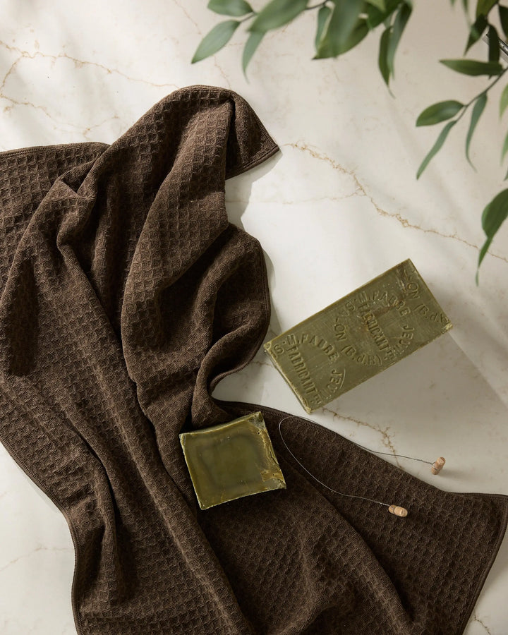 Espresso brown waffle-weave hand towel draped on white marble beside two bars of green olive-oil soap