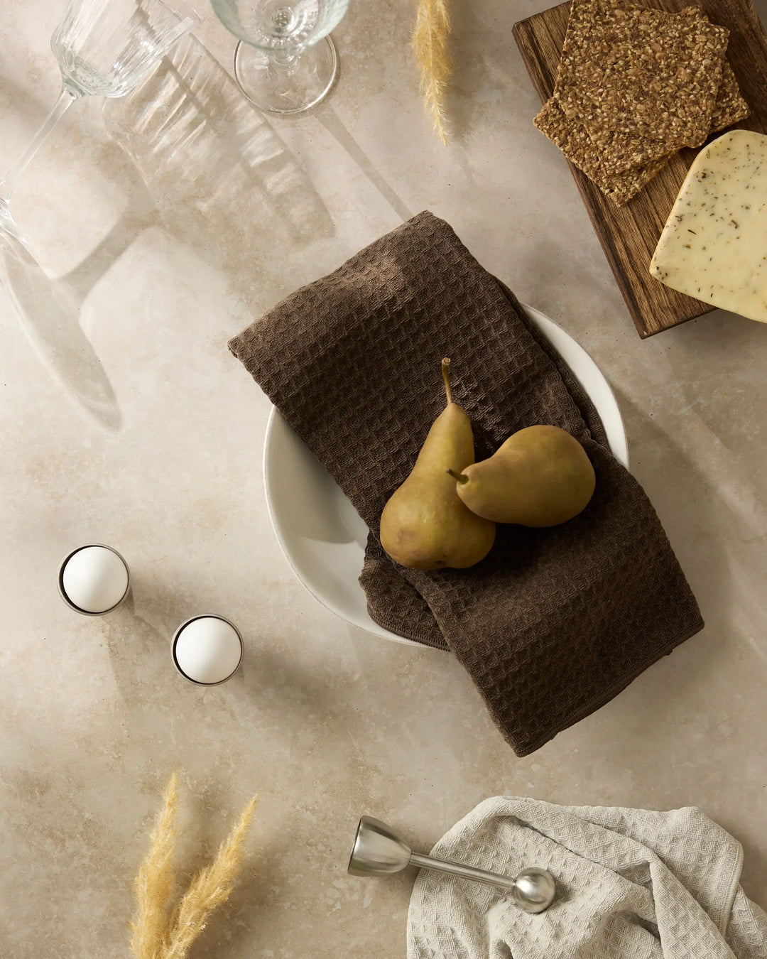 Espresso brown waffle-weave hand towel folded in a white bowl with two pears, on a table set with glassware and a cheese board