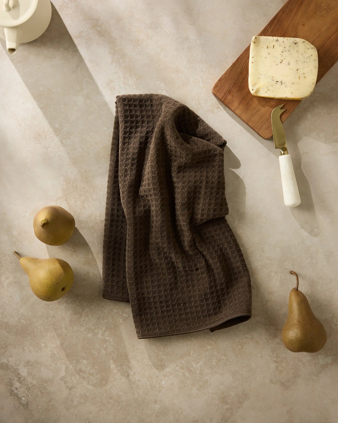 Espresso brown waffle-weave hand towel hanging in soft folds on a stone surface, with pears and a cheese board alongside