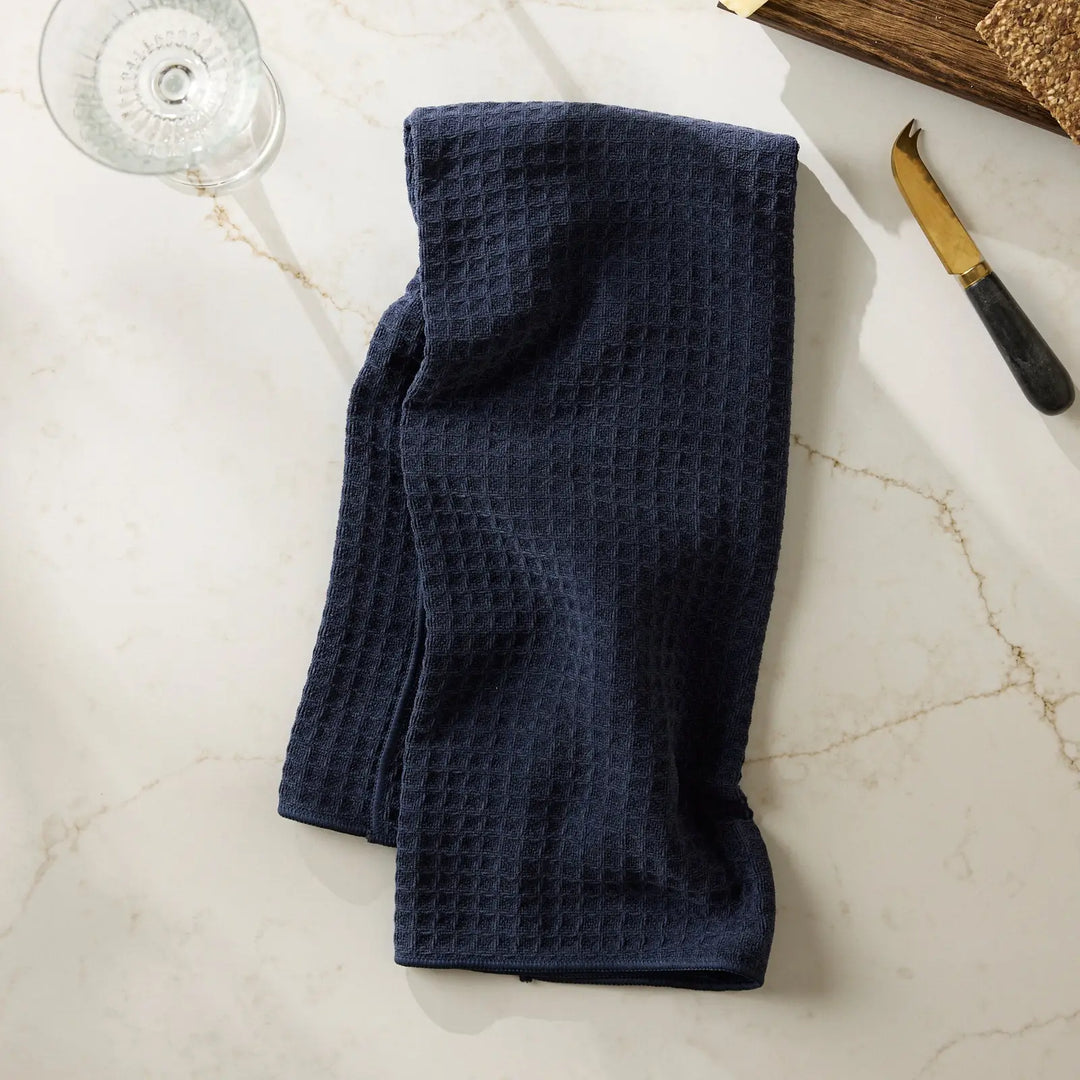 Navy blue waffle-weave hand towel folded on a white marble counter beside a wine glass and a brass cheese knife.