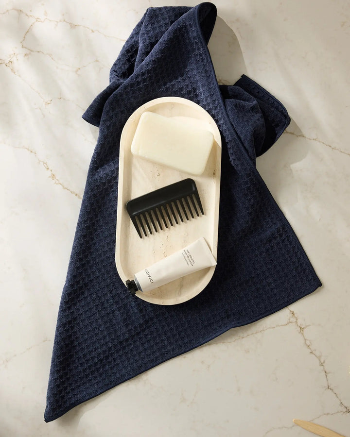 Navy blue waffle-weave hand towel spread on marble under a travertine tray holding a bar of soap, a black comb, and a tube of hand cream.