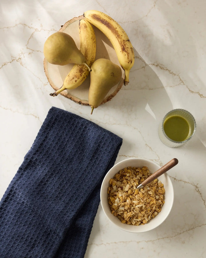 Navy blue waffle-weave hand towel folded on a marble counter beside a bowl of granola, a green smoothie, and a board of pears and bananas.