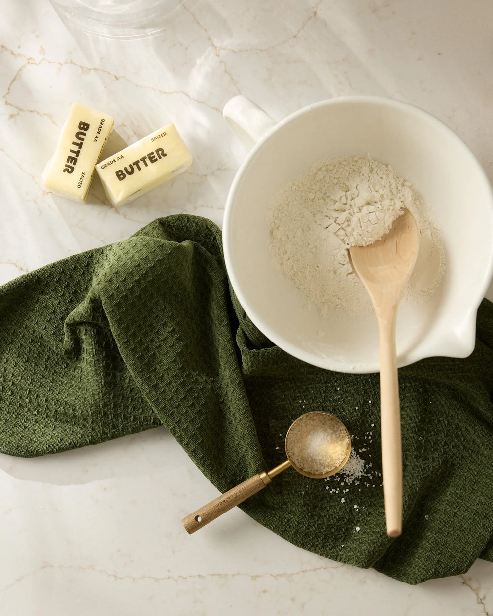 Matcha green waffle-weave hand towel under a white mixing bowl of flour, with a wooden spoon, sifter, and butter