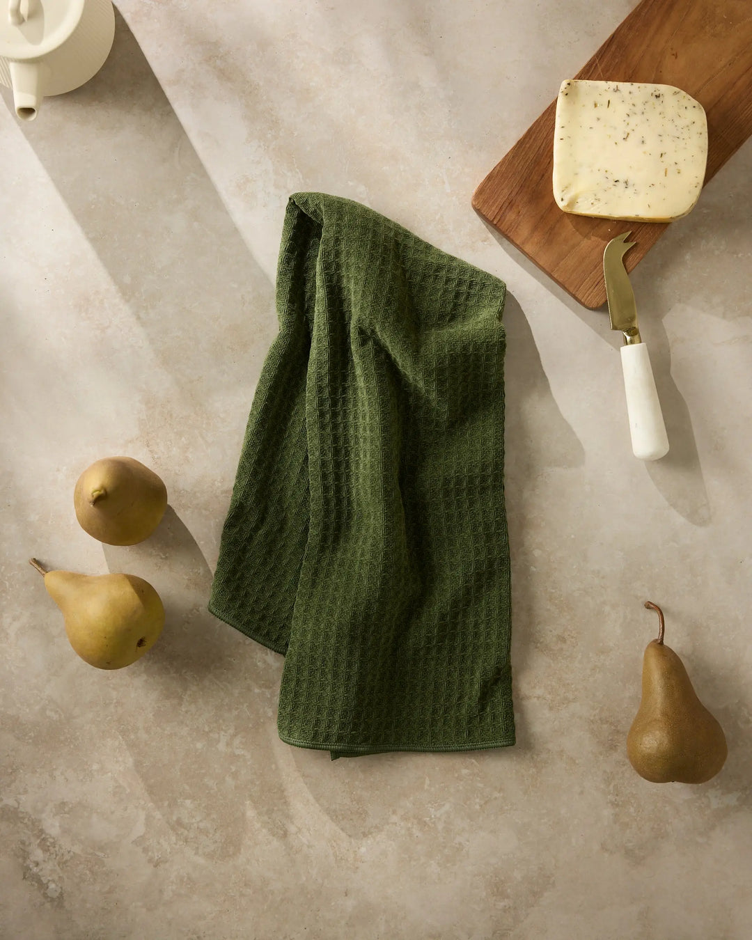 Matcha green waffle-weave hand towel hanging in soft folds on stone, with pears and a cheese board alongside
