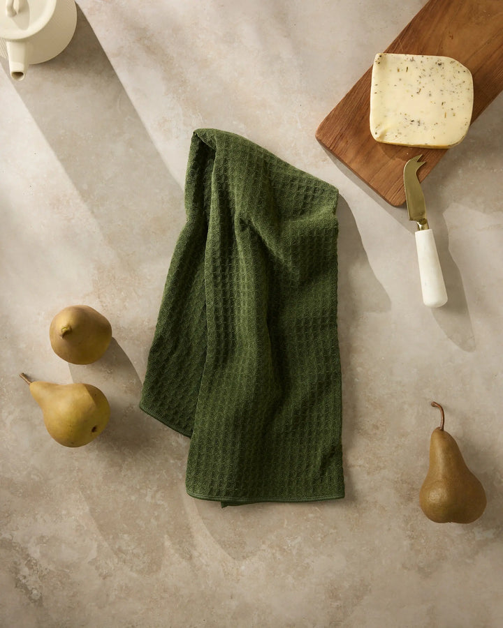 Matcha green waffle-weave hand towel hanging in soft folds on stone, with pears and a cheese board alongside
