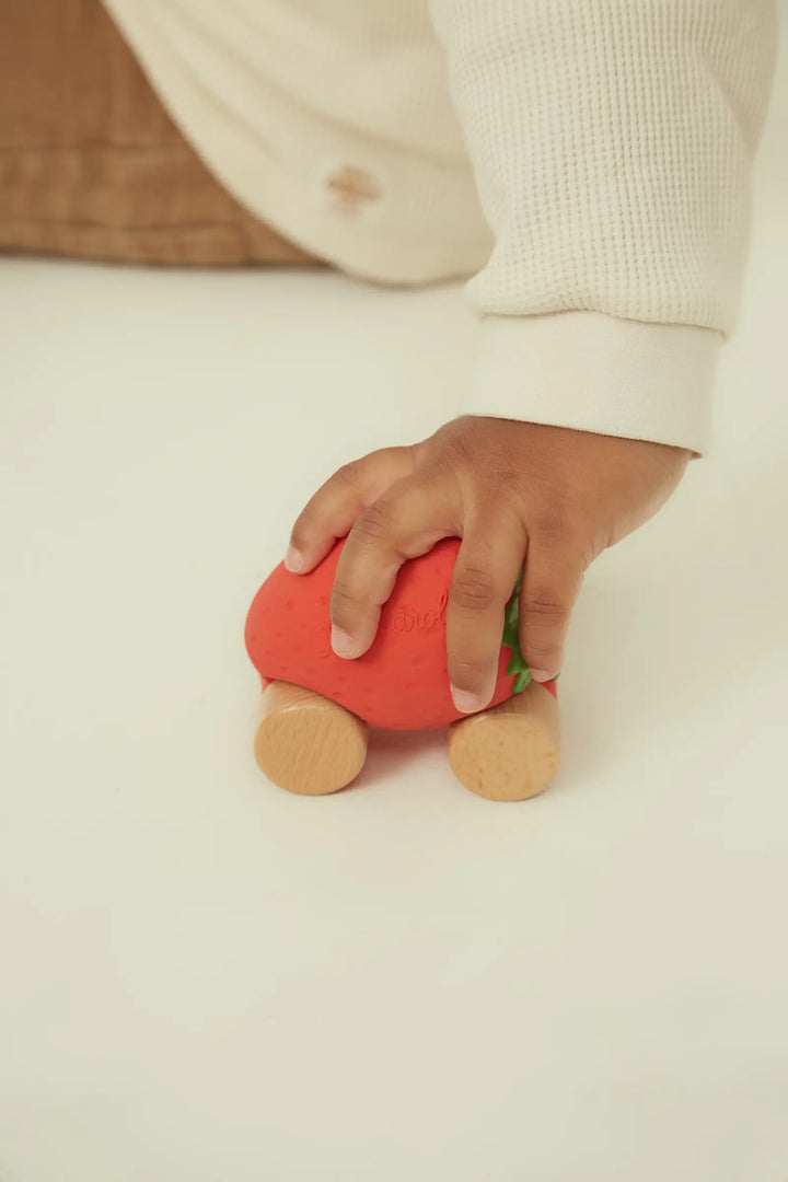 A hand resting on the red rubber strawberry car, showing its size against a pale background.