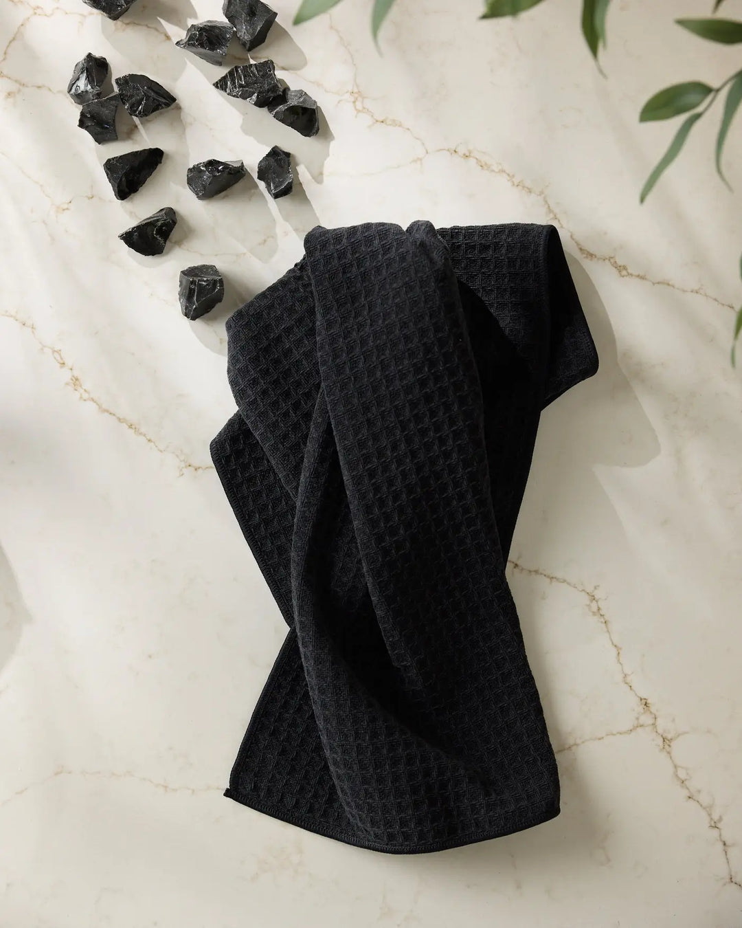 Black waffle-weave hand towel draped on cream marble beside chunks of raw obsidian and green eucalyptus leaves