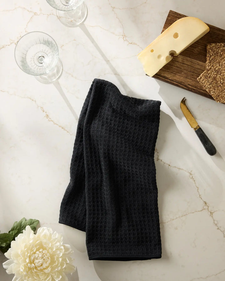Black waffle-weave hand towel folded on a marble counter with wine glasses, a cheese board, and a brass-handled cheese knife