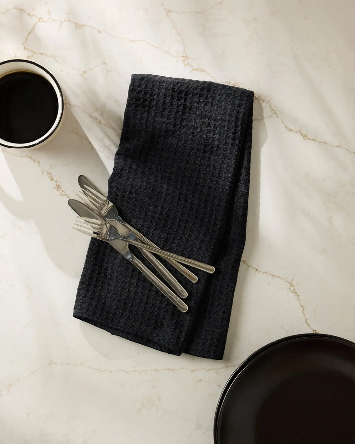 Black waffle-weave hand towel folded flat with silver flatware resting on it, beside a cup of black coffee and a dark plate