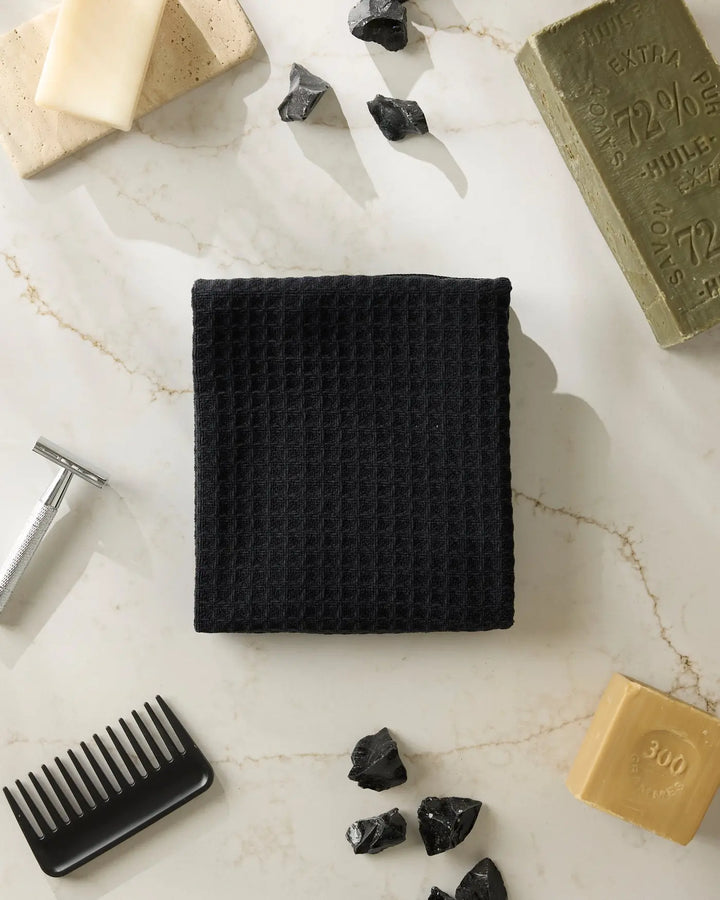 Black waffle-weave hand towel folded into a square on marble, surrounded by bar soap, a safety razor, a black comb, and obsidian pieces